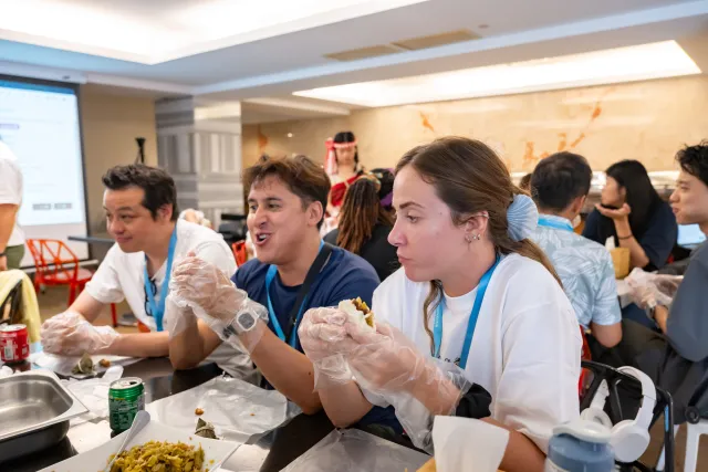 Participants wearing disposable gloves eat food together at a long table indoors, smiling and engaged