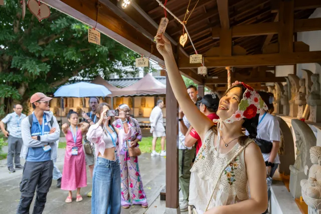 A smiling woman reaches up to hang a wooden wish plaque at a Japanese-style temple shrine as a group of visitors watches