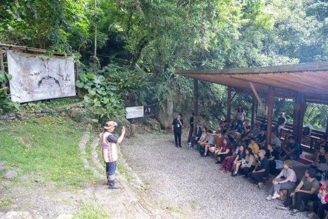 A guide in traditional dress leads an outdoor talk for a seated crowd under a forest shelter