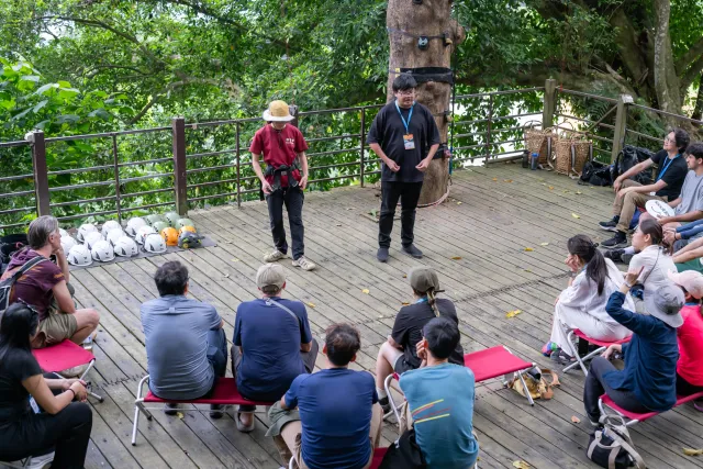 Two instructors brief seated participants on a forested wooden deck with helmets arranged nearby