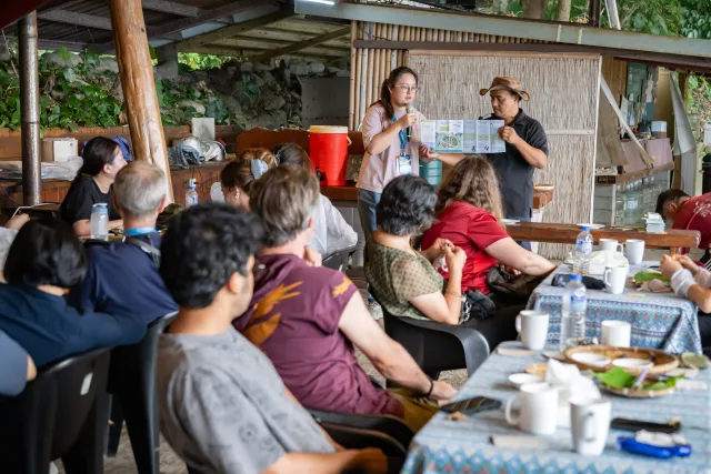 Two presenters address a seated group of participants in an open-air pavilion surrounded by greenery