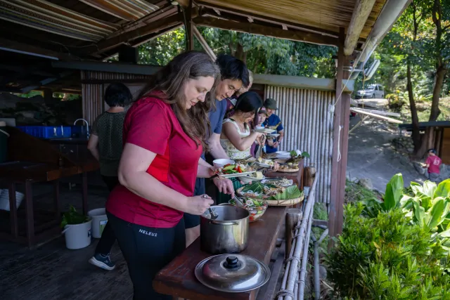 Participants serving themselves food along an outdoor covered counter surrounded by lush greenery