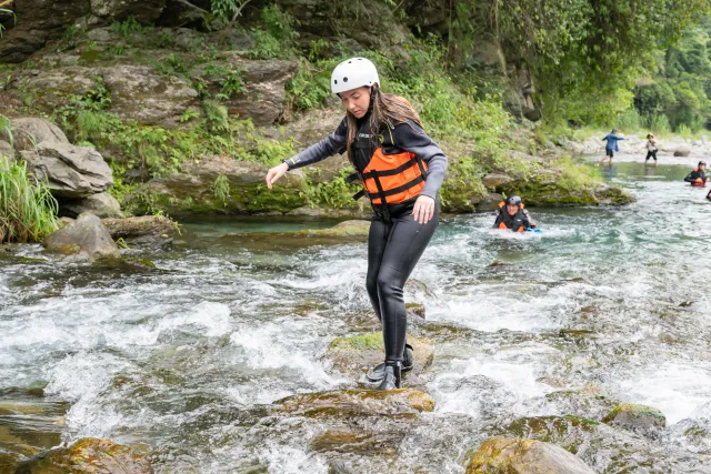 A participant in wetsuit and life jacket carefully steps across rocky river rapids