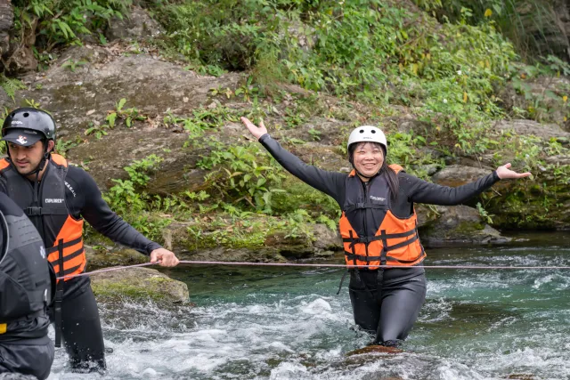 A smiling woman wades through a rocky river with arms outstretched, wearing a helmet and life jacket