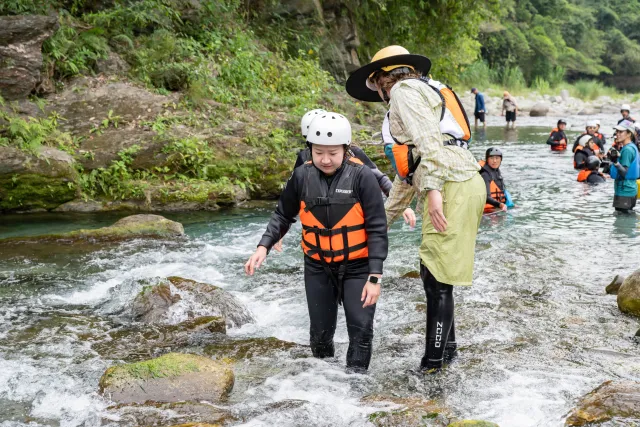 Guide in rain gear helps helmeted participant in life vest cross a rocky river