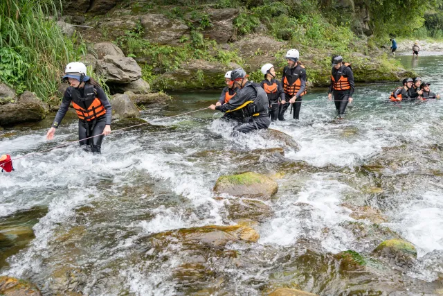 A group of participants in life jackets and helmets wading across a rocky rushing river
