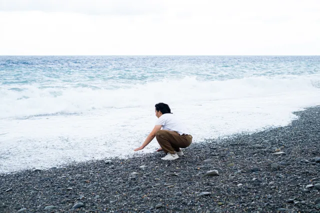 person crouching alone on a pebble beach, gazing at ocean waves