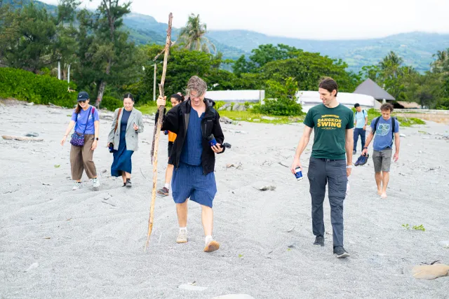 A group of visitors walks casually across a sandy beach toward the camera, with lush green hills in the background; one person carries a tall wooden stick.