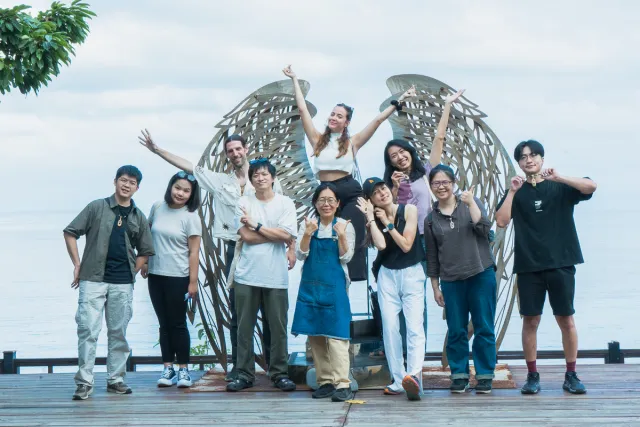 Group of ten people posing cheerfully in front of a large wing sculpture by the sea