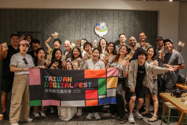 Large group of smiling participants poses indoors holding a Taiwan Digital Fest banner and small gift bags