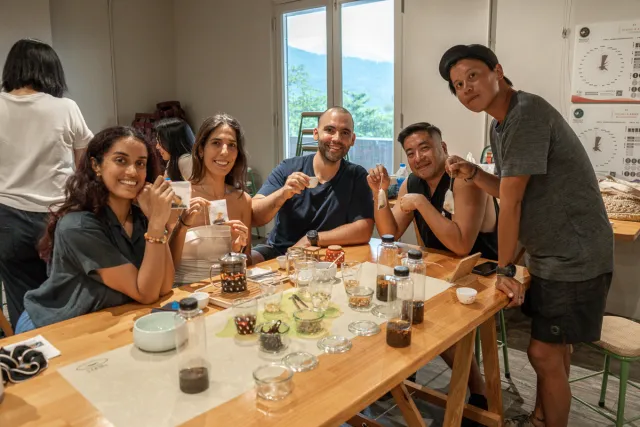 Five participants smile and hold up small cards at a tea and coffee tasting workshop indoors