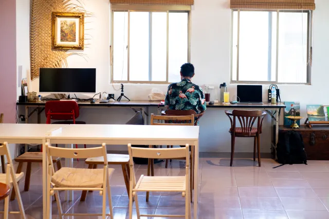 a person in a floral shirt sits alone working at a desk with monitors by bright windows