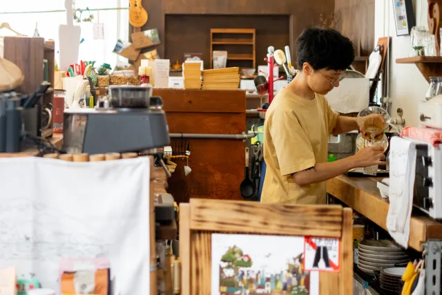 A person in a yellow T-shirt brews pour-over coffee at a cluttered wooden café counter