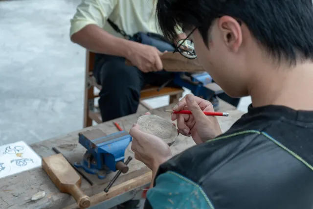 A young participant carefully carves a small wooden piece with a pencil at a workshop table
