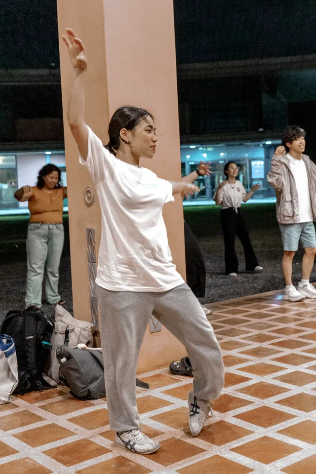 A woman in a white T-shirt and grey sweatpants dances hip-hop outdoors at night while others practice in the background