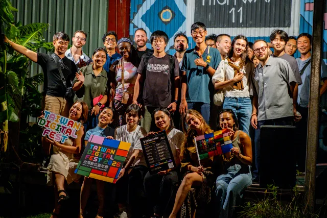 A diverse group of smiling attendees poses together outdoors at night holding Taiwan Digital Fest event programs