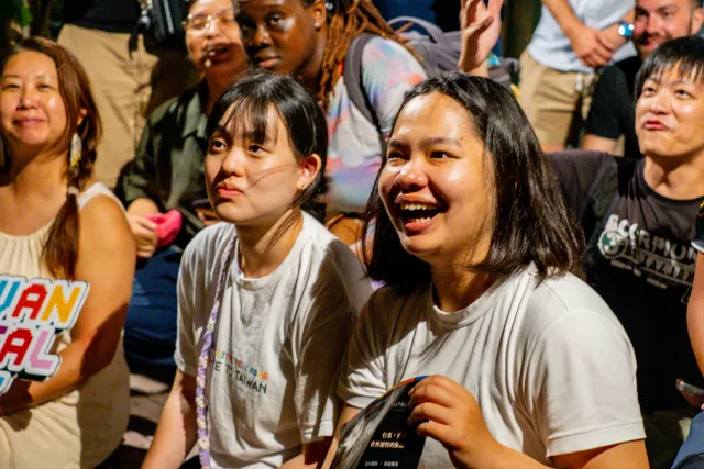 Two young women in white T-shirts laugh and smile while seated in a crowd at an evening event