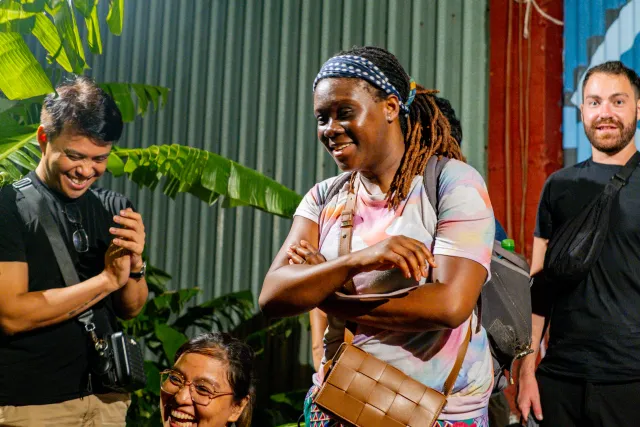A smiling woman with dreadlocks stands confidently outdoors at a night event, surrounded by laughing attendees