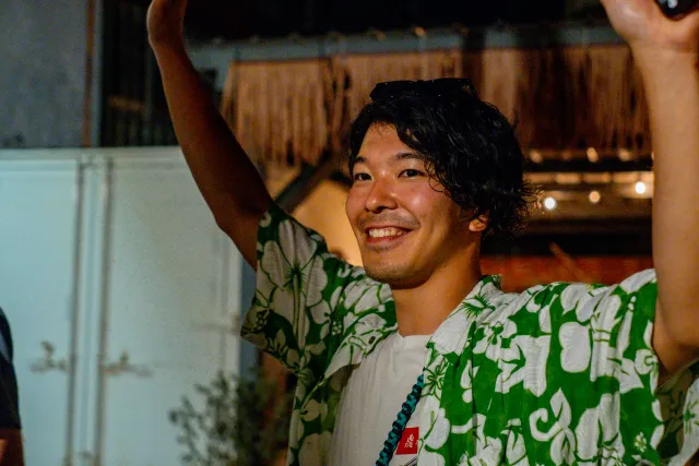 A smiling young man in a floral shirt raises his arm joyfully at an outdoor evening event