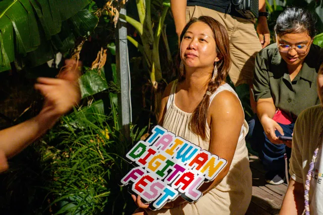 A woman holds a colorful Taiwan Digital Fest sign outdoors among tropical plants