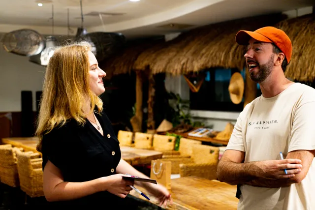 Two people laugh and chat warmly inside a venue with a thatched-roof structure