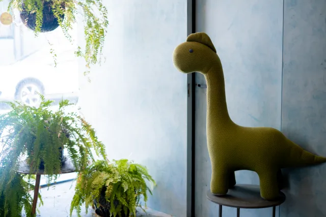 A knitted green dinosaur stuffed toy sits on a stool beside potted ferns near a window