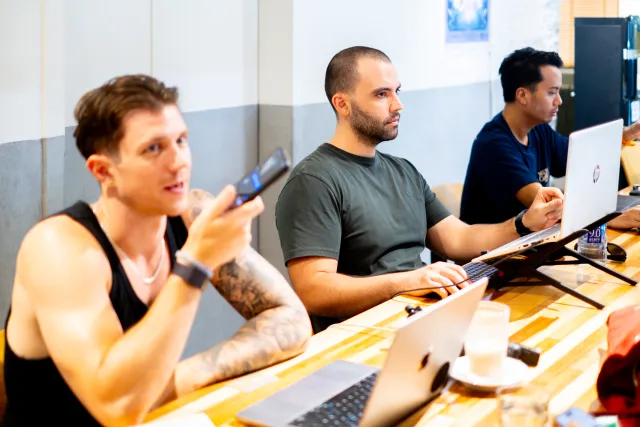 Three men sit at a long wooden table with laptops, attentively watching a presentation