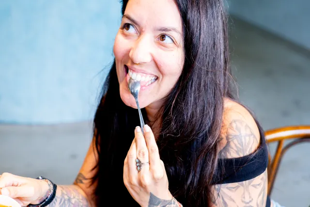 A tattooed woman smiles brightly while eating with a spoon at a cafe table