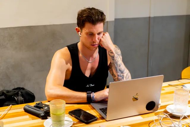 tattooed man in black tank top working intently on a laptop at a cafe table