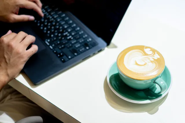 Hands typing on a laptop beside a latte art coffee cup on a white table
