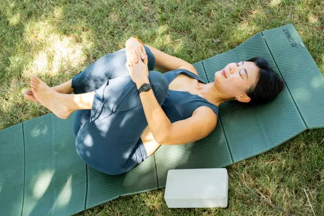A woman lies on a yoga mat outdoors on grass, eyes closed, hugging her knees in a supine stretch