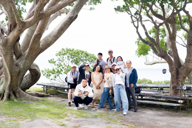 A group of about ten people posing together under large shade trees beside the ocean