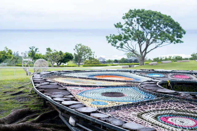 Colorful woven art installation on a boat hull with ocean and tree in background