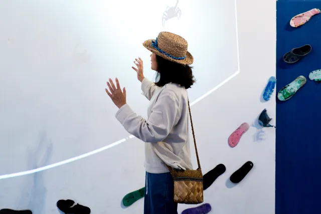A woman in a straw hat touches a white exhibition wall displaying flip-flops