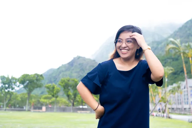 A smiling woman in a navy top stands outdoors with misty mountains and palm trees behind her