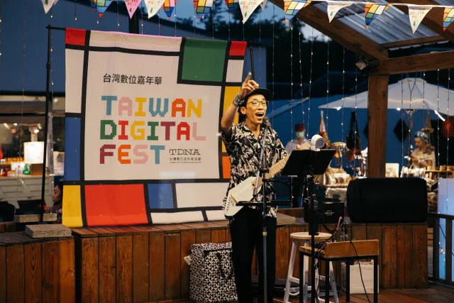 A speaker gestures enthusiastically on an outdoor stage in front of a Taiwan Digital Fest banner at dusk