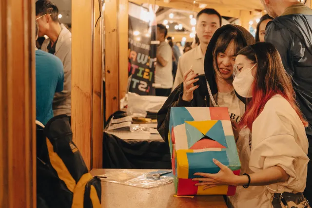 Two young women examine a colorful geometric paper lantern at a night market booth