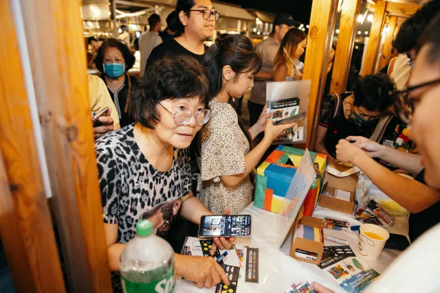 Elderly woman and younger visitors browse printed materials at an outdoor night market booth