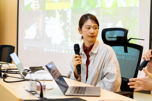 A woman speaks into a microphone at a conference table with laptops and a projected screen behind her