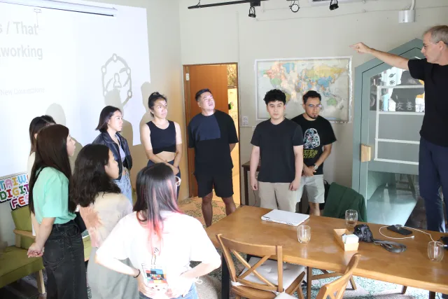 A speaker gestures while addressing a small group of attentive attendees in a casual indoor venue