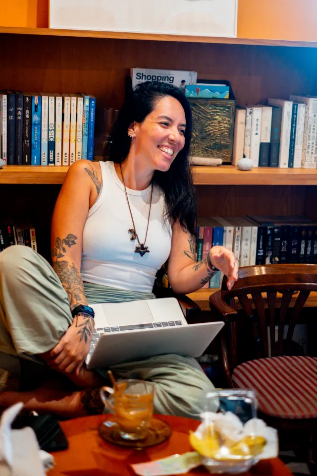 A tattooed woman smiles broadly while sitting with a laptop in a warm bookshelf-lined cafe