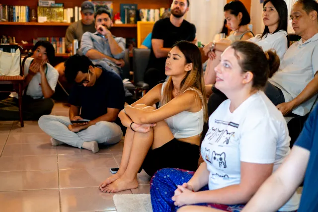 Audience members sitting on the floor and chairs attentively watching a speaker in a cozy indoor venue