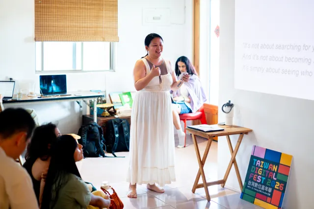 A woman in a white dress speaks to a seated indoor audience beside a projected slide