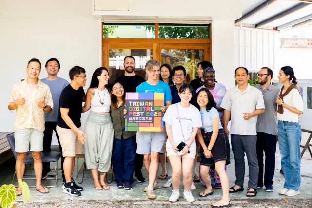 A diverse group of about fifteen people posing cheerfully outdoors in front of a white building, holding a Taiwan Digital Fest 2026 sign