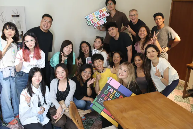 A large diverse group of smiling people poses together indoors holding Taiwan Digital Fest signs