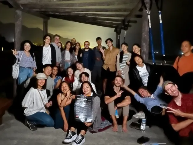 A large group of smiling participants poses together outdoors at night under a wooden shelter
