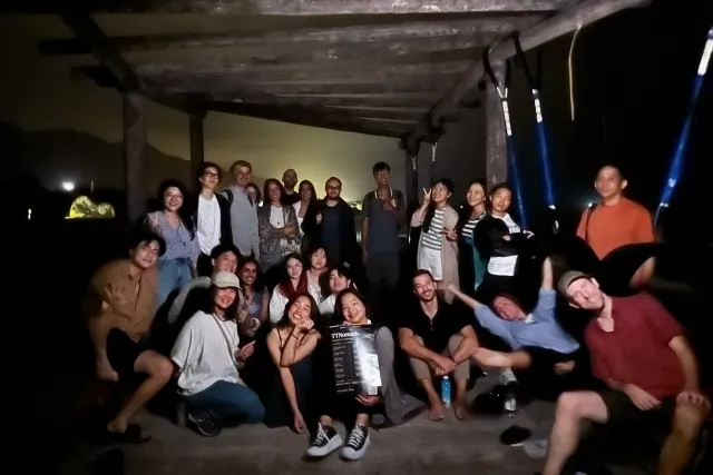A large group of people pose together for a photo indoors at night, smiling and gesturing