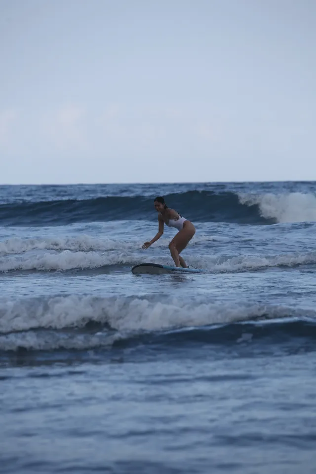 A woman in a white swimsuit balances on a surfboard riding a small wave