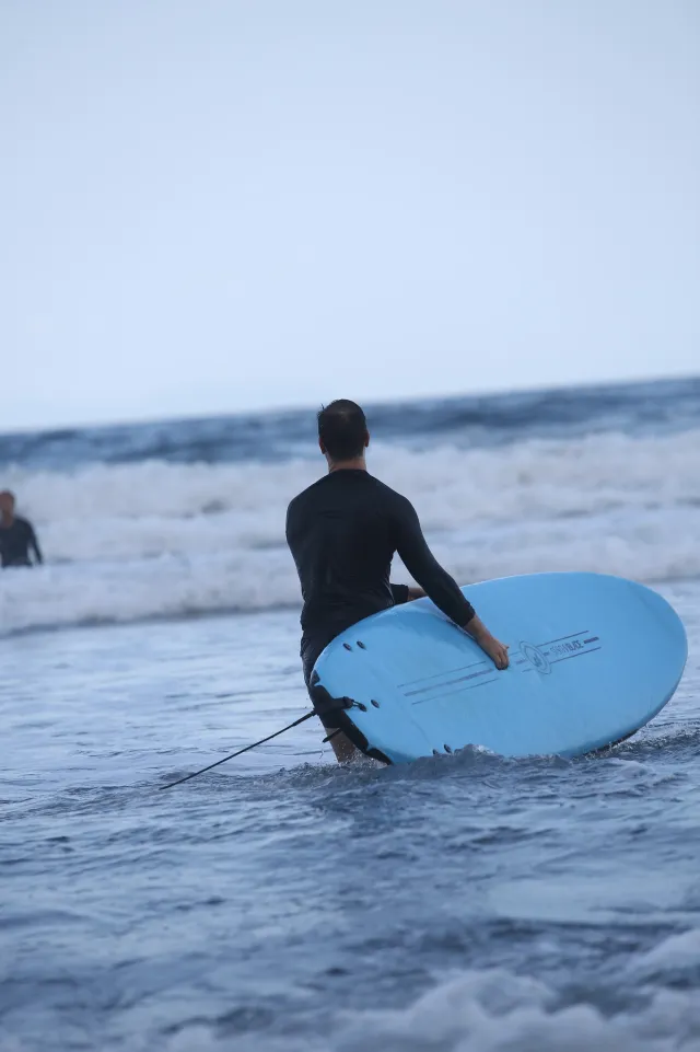 A surfer in a black wetsuit wades into the ocean carrying a blue foam board