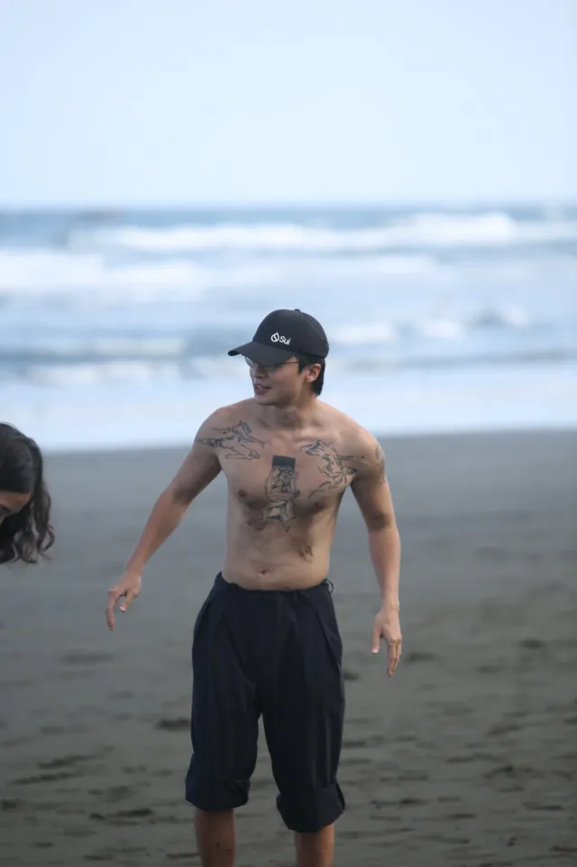 Tattooed shirtless man in a cap smiling on a dark sand beach with waves behind him